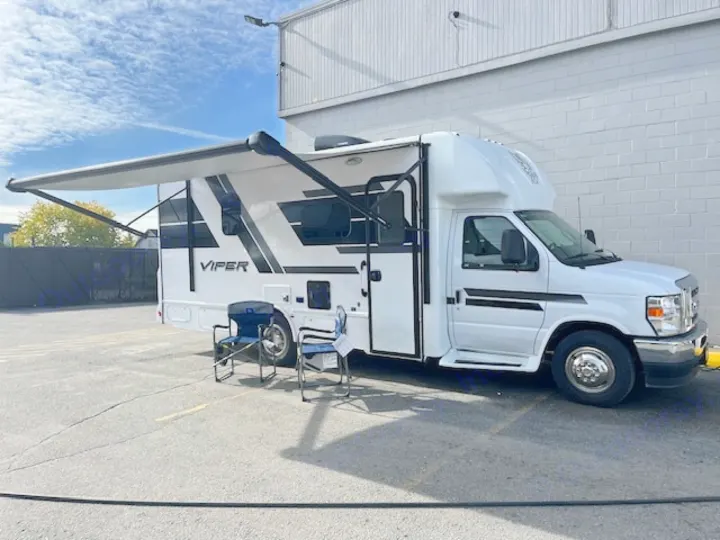 A white Viper RV rental with an extended awning and blue chairs sits on asphalt next to a building, ready for adventure.. Nexus Viper 2026