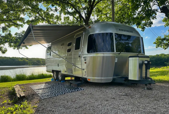 Serene Airstream RV rental camping by a sunny lake, nestled under shady trees for a perfect nature escape.. Airstream Flying Cloud 2012