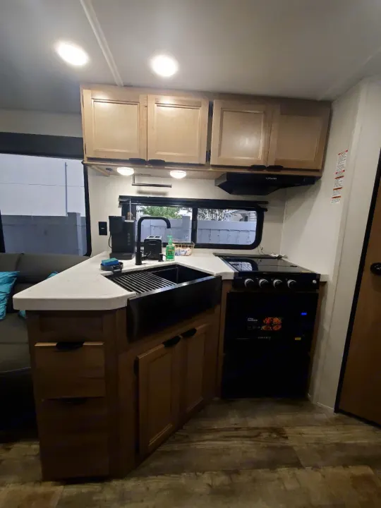 Inside an RV rental, a compact kitchen features light wood cabinets, a black sink, stove, and white countertops for travel.. Forest River WOLF DEN 2024