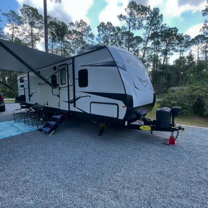 This 2022 rv rental shows a white travel trailer with an awning extended, parked on gravel at a campsite with pine trees.. Keystone RV Bullet Ultra Lite 2022