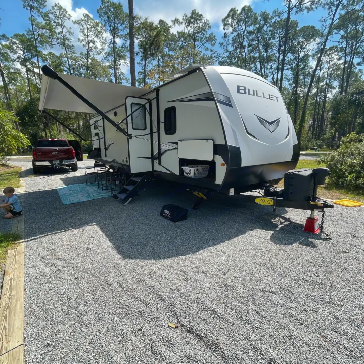 Camping with an RV rental! A Bullet travel trailer set up at a gravel campsite among pine trees, with a truck and child nearby.. Keystone RV Bullet Ultra Lite 2022