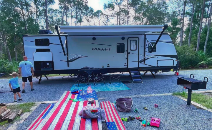 Family camping with their RV rental Bullet travel trailer, featuring an extended awning, picnic setup, and grill at a serene campsite.. Keystone RV Bullet Ultra Lite 2022