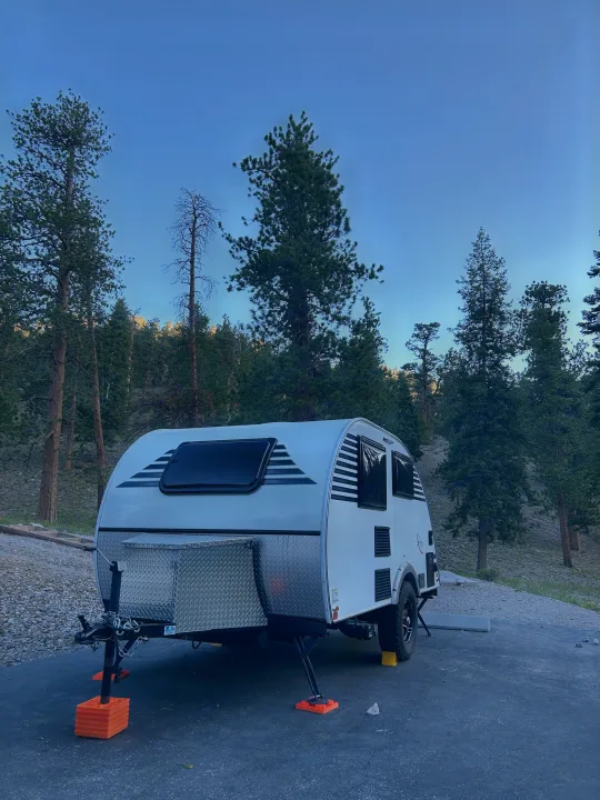This teardrop RV rental, a compact camper, is set up for a serene camping trip among tall pine trees under a clear sky.. Xtreme Outdoors Xtreme Outdoors Trailer 2022