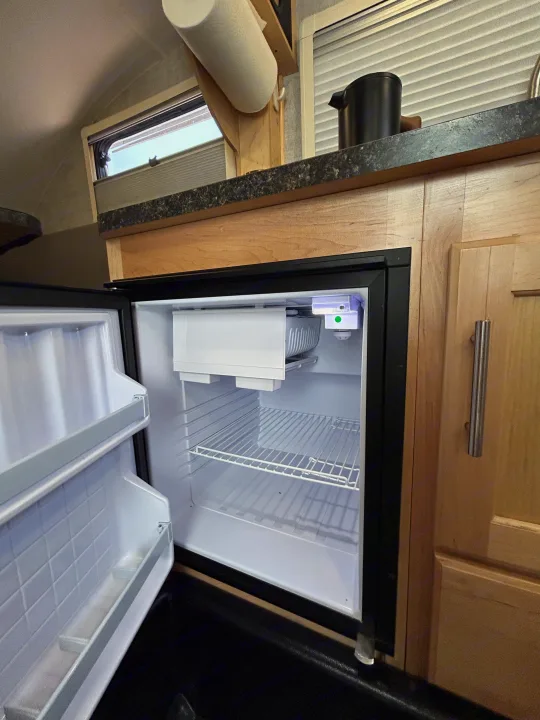 Open, empty mini-fridge in an RV rental, showing compact kitchen with counter, cabinet, and window.. Xtreme Outdoors Xtreme Outdoors Trailer 2022