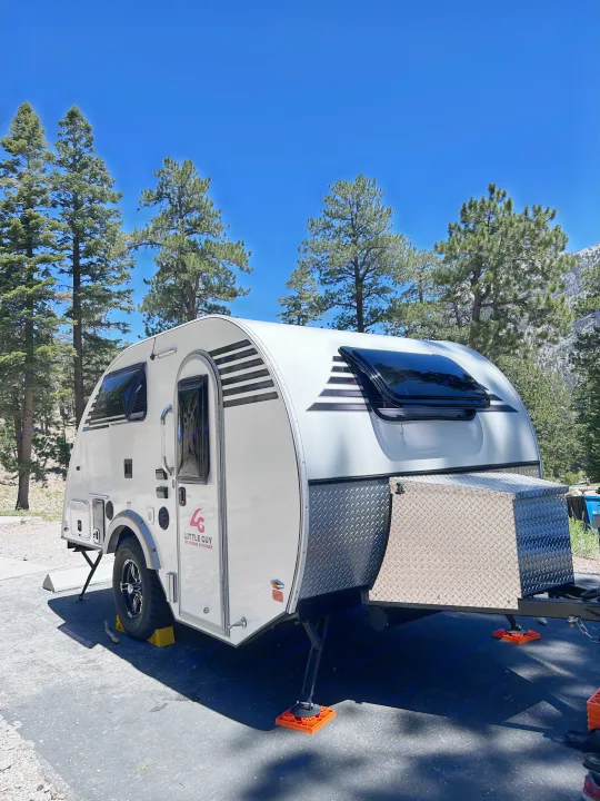White teardrop RV rental parked amidst pine trees under a clear blue sky, featuring black and white striped accents. Ideal for camping.. Xtreme Outdoors Xtreme Outdoors Trailer 2022