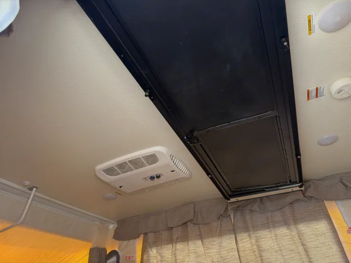 The interior ceiling of an RV rental pop-up camper, featuring an AC unit, ceiling lights, a black folding bed panel, and safety warning labels.. Forest River Flagstaff M.A.C. 2022