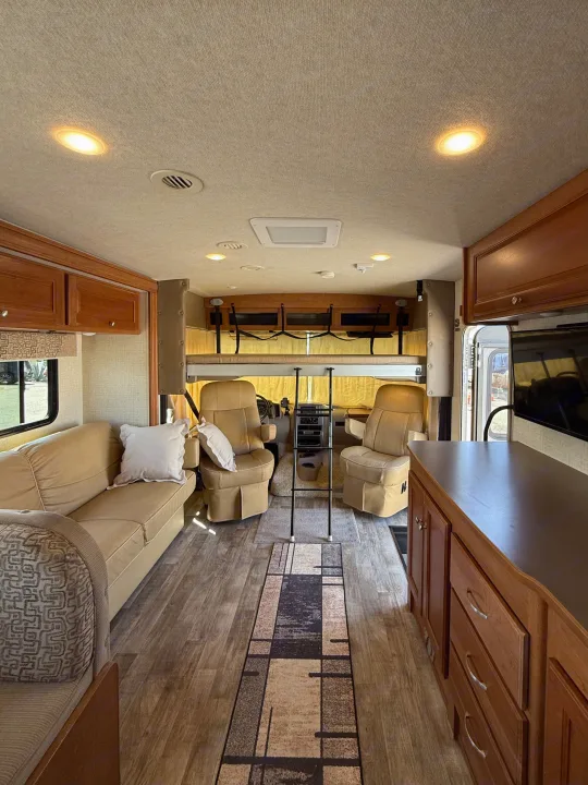 Comfortable RV rental interior featuring a sofa, captain's chairs, overhead bunk, and wood-look flooring with a runner.. Winnebago Vista 2017