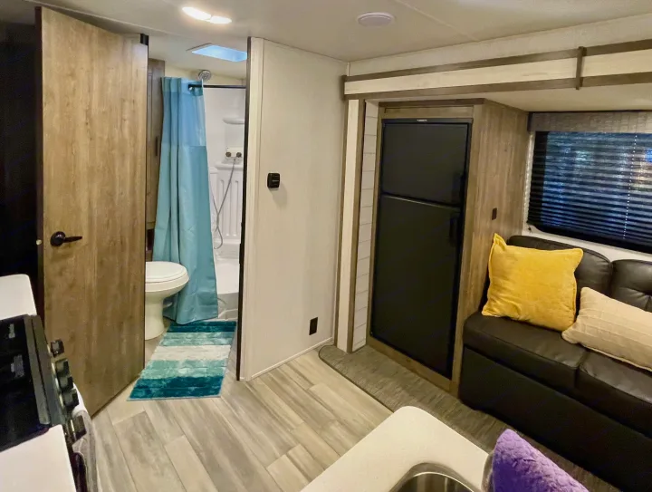 This inviting RV rental interior features a bathroom with a shower and toilet, a kitchen counter, a black refrigerator, and a cozy sofa with pillows by a window.. Heartland RVs Pioneer 2023