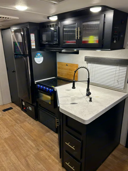 Dark-themed RV rental kitchen: black fridge, oven, microwave, sink with gooseneck faucet, marbled counter, and wood floor.. Forest River Cherokee 2021