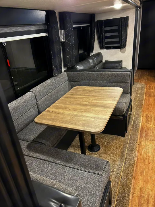 Modern grey RV rental dinette featuring U-shaped booth seating, a light wood table, and dark curtains. A cozy travel space.. Forest River Cherokee 2021