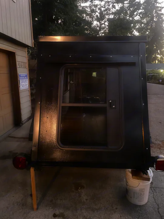 Rear view of a compact black custom-built camper with a windowed door, propped on blocks, ready for **rv rental**.. Custom Squaredrop Trailer 2020