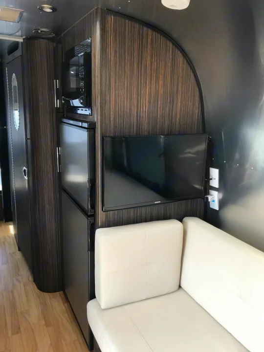 This modern **rv rental** interior features dark wood cabinets, a microwave, fridge, wall TV, and a light sofa.. Airstream International 2018