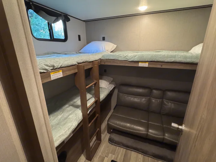 RV rental bunk room: three single bunk beds with soft green blankets, a ladder, and a leather-look gray sofa.. Crossroads RV Zinger 2020