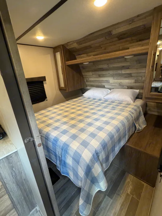 This cozy rv rental bedroom features a blue plaid quilt, white pillows, a wood-look headboard, and overhead storage cabinets.. Crossroads RV Zinger 2020