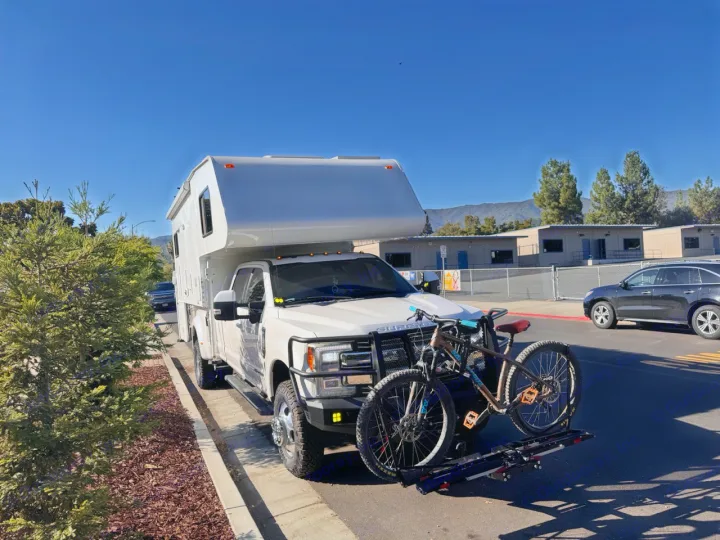 White truck camper RV rental, equipped with bikes, parked on a sunny street, ready for outdoor adventures.. Ford F350 Diesel Double Cab 2017