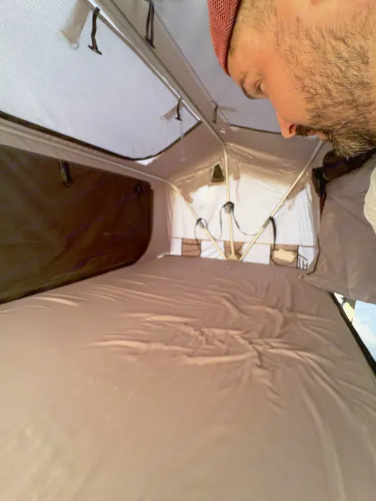Inside an RV rental's pop-up tent, showing a comfortable sleeping area with a neatly made bed and storage pockets. A man peers in from above.. Silver Eagle Manufacturing M1102 2013