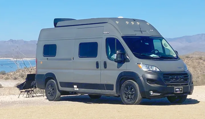 This grey camper van RV rental is parked by a lake, with desert mountains in the background and folding chairs visible nearby.. Winnebago Solis Ram 2025