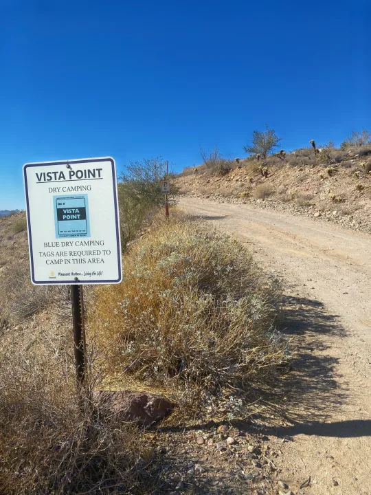 Desert scene with a sign for Vista Point dry camping, indicating RV rental tags are required. A dirt road winds into the distance.. Winnebago Solis Ram 2025