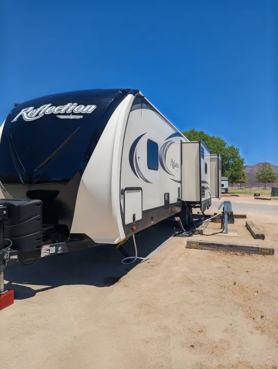 A "Reflection" RV rental is set up at a sunny campsite, showing slides out, hookups, and mountains in the background.. Grand Design Reflection 2019