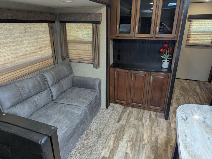 Cozy RV rental living space featuring a comfy grey couch, dark wood cabinetry, and multiple windows with blinds.. Grand Design Reflection 2019
