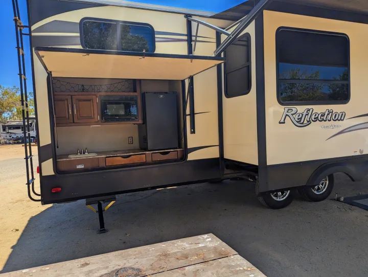Cream-colored Reflection rv rental showcasing an open outdoor kitchen with a sink, microwave, and fridge at a sunny campsite.. Grand Design Reflection 2019
