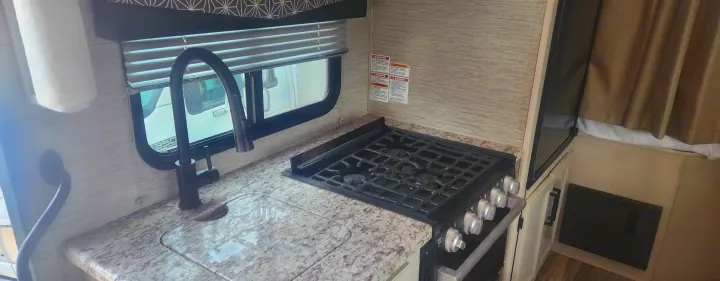 RV rental kitchen: modern black faucet, sink, and gas stove on a granite-look counter with a window and blinds.. Thor freedom elite 2022