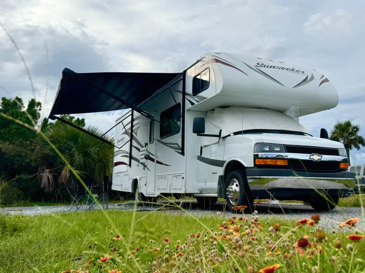 This Sunseeker RV rental is parked at a serene campsite with its awning extended, surrounded by wildflowers and lush greenery.. Forest River Sunseeker 2017