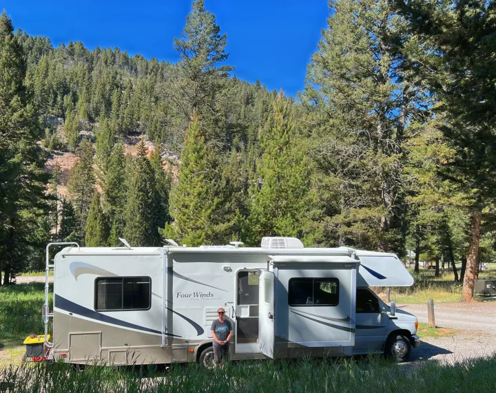 Woman next to an RV rental in a sunny mountain forest. The grey and white RV is parked among tall pine trees.. Four Winds 5000 2006