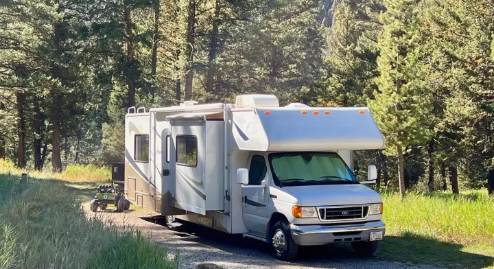 This white RV rental is parked on a gravel path in a sunny pine forest, ready for camping. A green child's ride-on toy is visible.. Four Winds 5000 2006