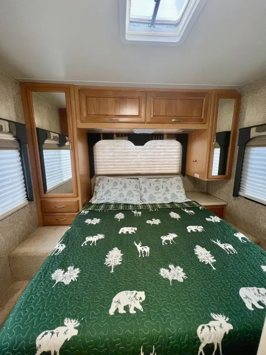 An RV rental bedroom features a green quilt with forest animals & trees, flanked by wood cabinets & mirrors.. Four Winds 5000 2006