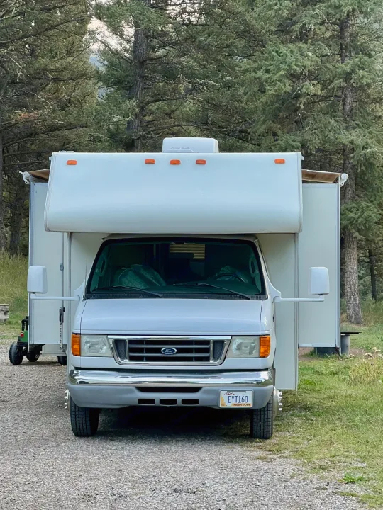 A white RV rental, with a Montana license plate, is parked among tall evergreen trees in a scenic outdoor campground.. Four Winds 5000 2006