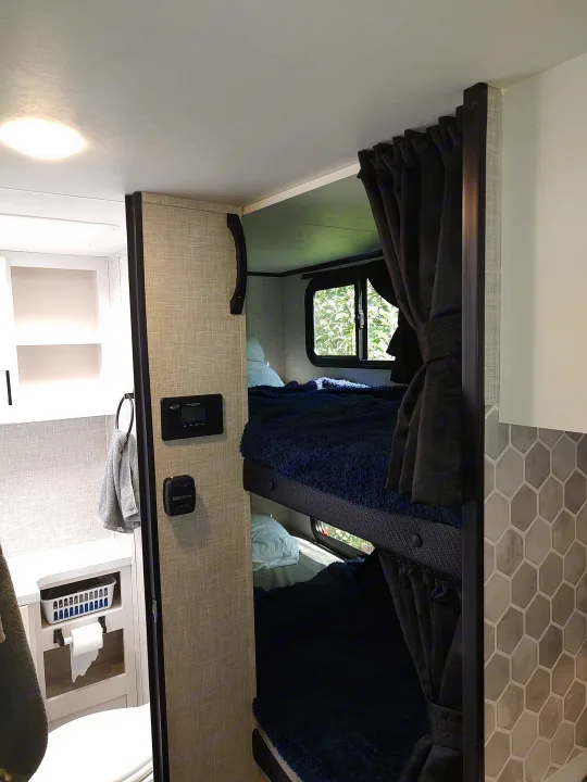 RV rental interior: cozy bunk beds with dark curtains, control panels, and a compact bathroom with hexagonal tiles.. Jayco Jay Feather Air 18mbh 2024