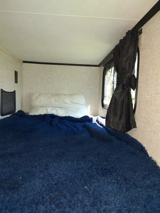 Inside an RV rental: a snug sleeping nook with a dark blue fluffy blanket, white pillow, and window with a tied black curtain.. Jayco Jay Feather Air 18mbh 2024