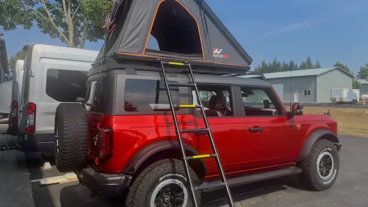 Red Bronco with a Naturhike rooftop tent and ladder, parked beside an RV rental van, ready for off-road adventure.. Ford Bronco Badlands 2023