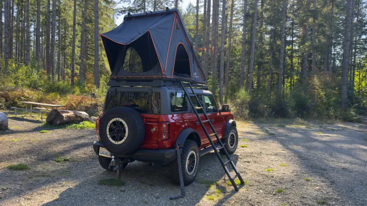 An rv rental alternative: Red Ford Bronco with rooftop tent set up for camping in a scenic forest clearing.. Ford Bronco Badlands 2023