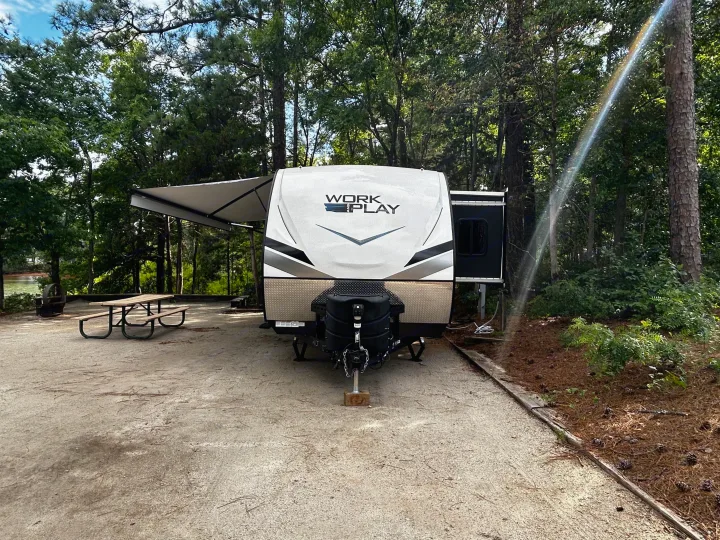 A "WORK AND PLAY" RV rental trailer is parked at a wooded campsite with an awning, picnic table, and distant lake.. Work and Play Toy Hauler 2022