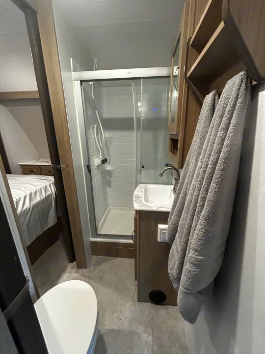 A functional rv rental bathroom featuring a shower, sink, toilet, and a bed seen through the doorway.. Work and Play Toy Hauler 2022