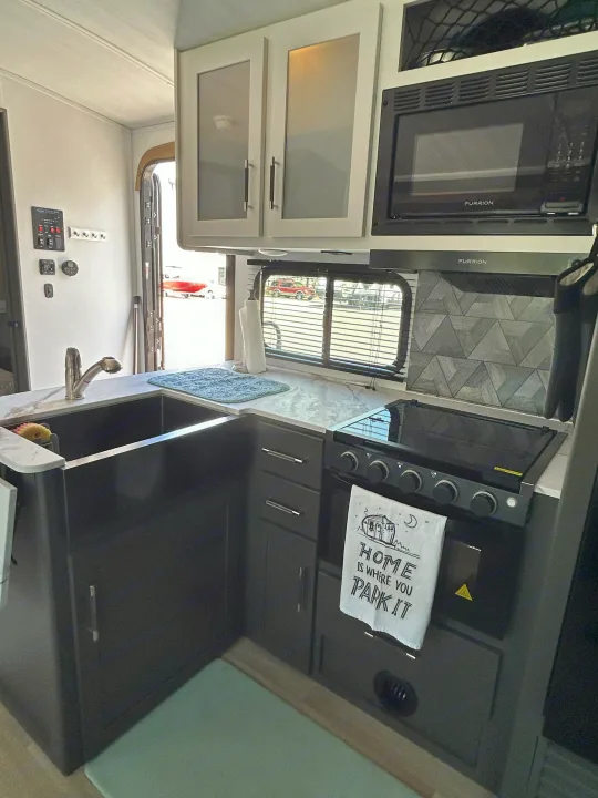 Compact rv rental kitchen featuring a dark sink, stove, microwave, light countertops, and grey cabinets.. Keystone RV Bullet Crossfire 2025