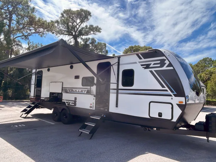 A modern white Bullet travel trailer rv rental, shown parked with an extended awning and entry steps, under a blue sky.. Keystone RV Bullet Crossfire 2025