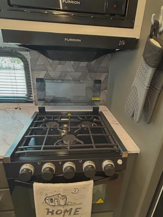 RV rental kitchen features: Black stove/oven, microwave, range hood, geometric backsplash, and hanging oven mitts.. Keystone RV Bullet Crossfire 2025