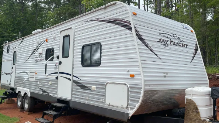 Jay Flight travel trailer RV rental, silver and white, parked in a wooded area, showing doors, windows, and dual axles.. Jayco Jay Flight 2016