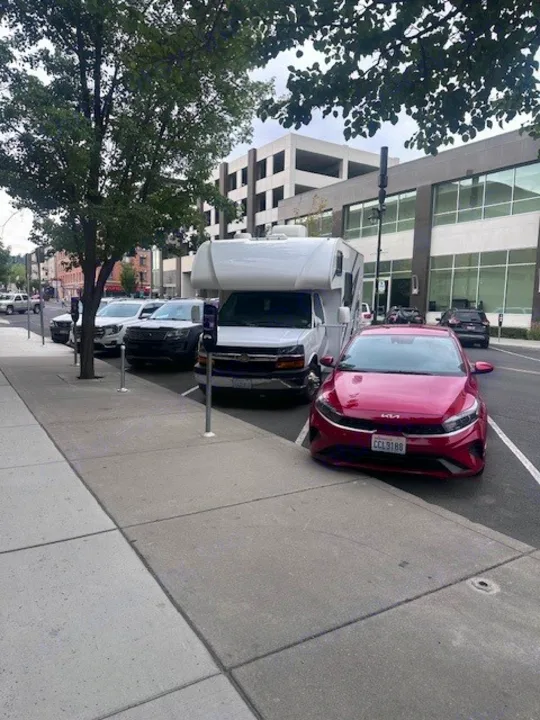 A red car and a white RV rental are street-parked in a city, with other vehicles and modern buildings in the background.. Thor Freedom Elite 2021