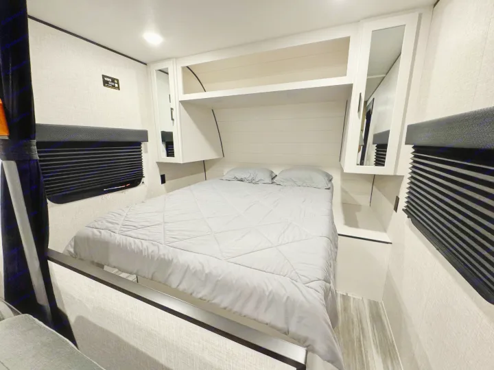 RV rental bedroom with a made bed, white overhead cabinets, and black pleated window blinds.. Jayco 260BH 2026