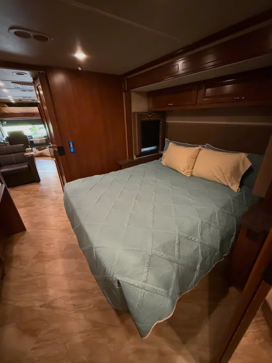 This inviting RV rental bedroom features a cozy bed with a light blue quilt, wooden cabinetry, and a glimpse into the living area.. Fleetwood Bounder 2016