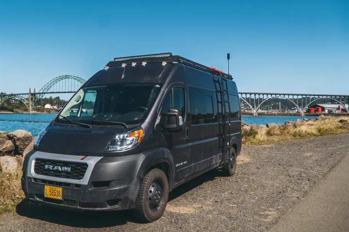 Black Ram ProMaster camper van, likely an RV rental, parked on a gravel road by a scenic waterfront with a large bridge.. Dave & Matt Vans Dave & Matt Vans Class B 2022