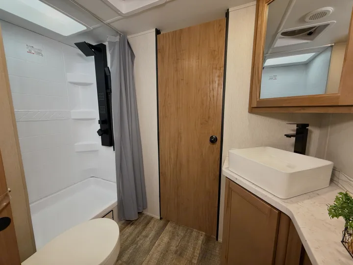 RV rental bathroom featuring a white shower enclosure, toilet, wooden door, and vanity with a modern vessel sink and black faucet.. Forest River Black Label 2024