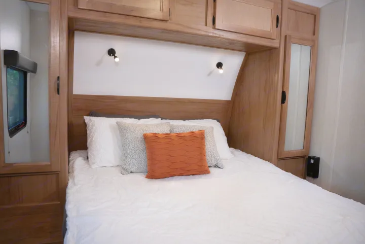 Interior view of an RV rental showing a bed with white bedding, accent pillows, wood cabinets, and reading lamps.. Forest River Black Label 2024