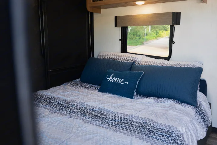 RV rental bedroom with stylish geometric bedding, blue pillows (one with 'home'), and a window looking out to the road.. Forest River Black Label 2024
