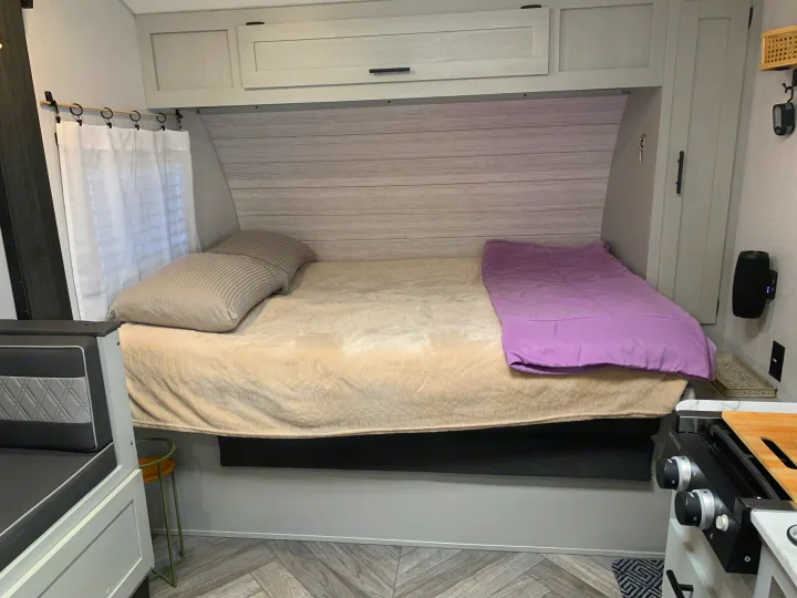 Cozy RV rental bedroom with a bed, striped pillows, lavender comforter, modern cabinetry, and a partial kitchen view.. Forest River Wildwood FSX Plantium 2022
