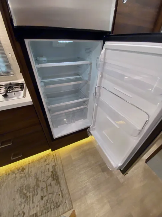 Open, empty RV rental refrigerator in a compact kitchen, with clear shelves and door storage, ready for groceries.. Winnebago View 2020